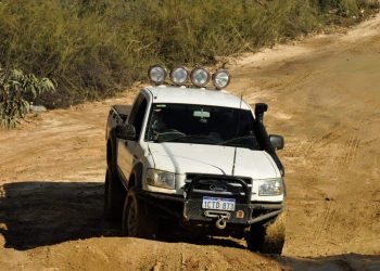 A white Ford pickup truck equipped with roof-mounted off-road spotlights and a bull bar, driving on a dirt road after spotlight installation.