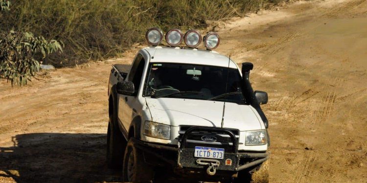 A white Ford pickup truck equipped with roof-mounted off-road spotlights and a bull bar, driving on a dirt road after spotlight installation.