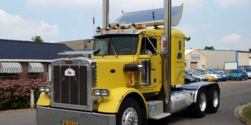 A bright yellow Peterbilt semi-truck parked on the road, showcasing chrome exhaust stacks, aerodynamic roof deflectors, and a powerful front grille design.
