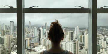 A woman standing by a large window looking out at a modern city skyline on a cloudy day.